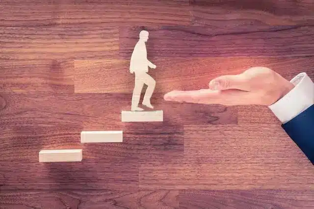 Een houten figuur die stappen omhoog zet richting een ondersteunende hand, symbool voor de ondersteuning die de expertise van bedrijfsarts Van Bennekom biedt